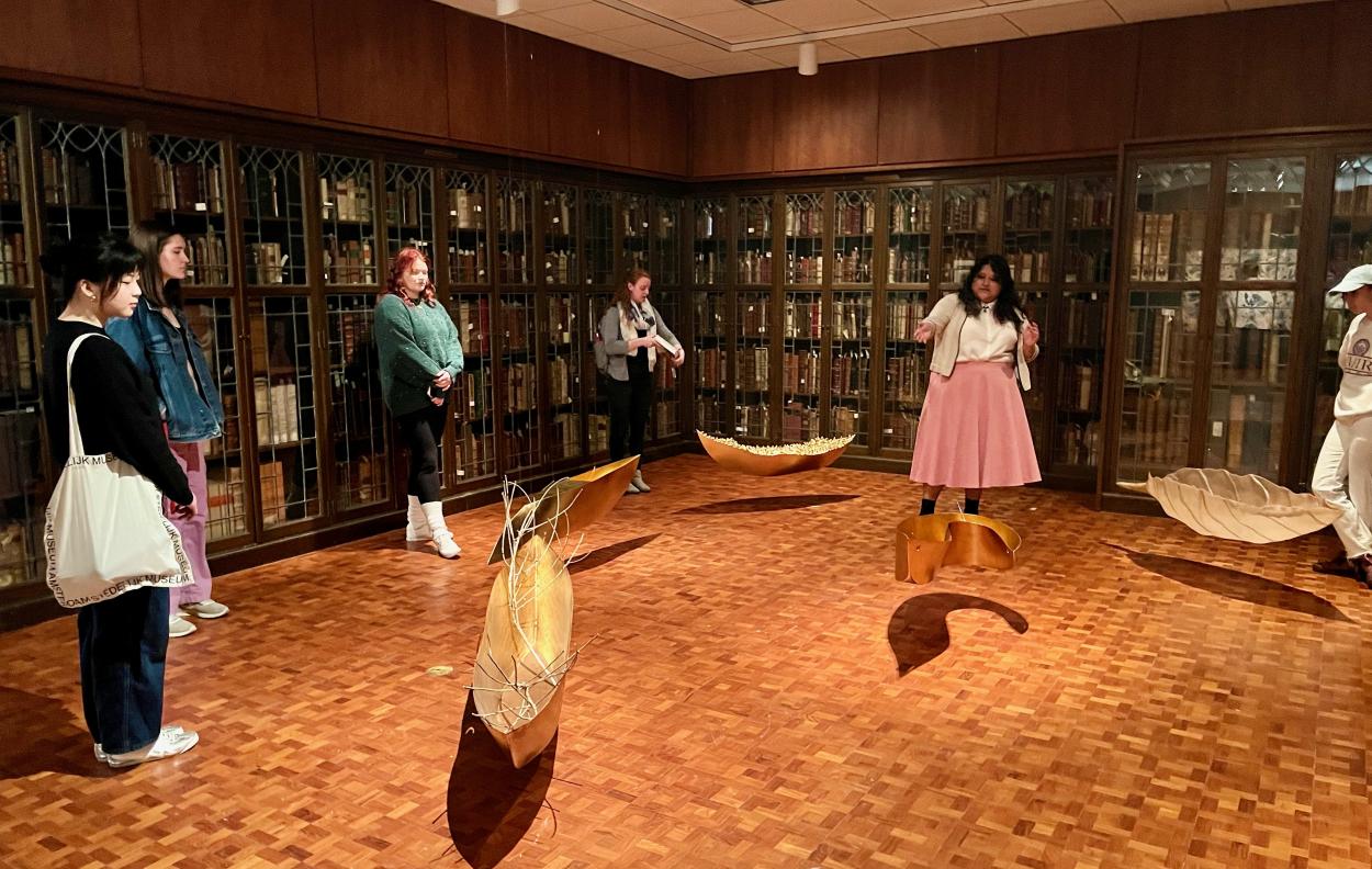 Joselyn Garcia leads a Friday Finds tour group through the Cecilia Paredes exhibition; she points to the gilded boat sculptures hanging in the gallery