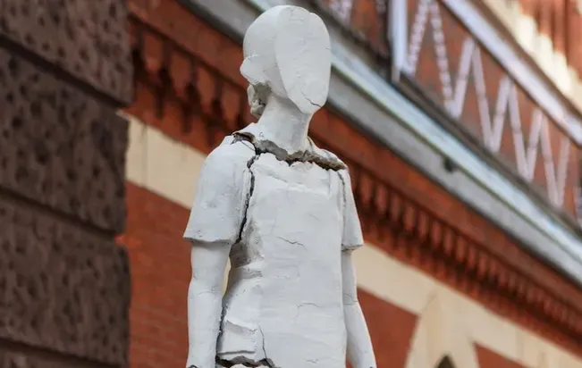 A photograph of Tania Bruguera's Monument to New Immigrants. The figure of a girl, faceless and cracking at seams, holds a teddy bear and looks expectantly forward.