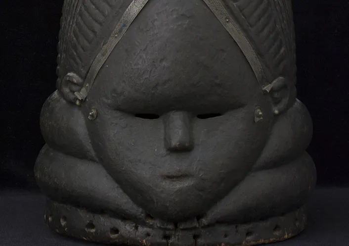 Blackened wooden mask of a diamond-shaped face, framed by a triangular hairline and chinline. The hairline is embellished with applied metal strips, above which is an elaborate coiffure of braids and bun.