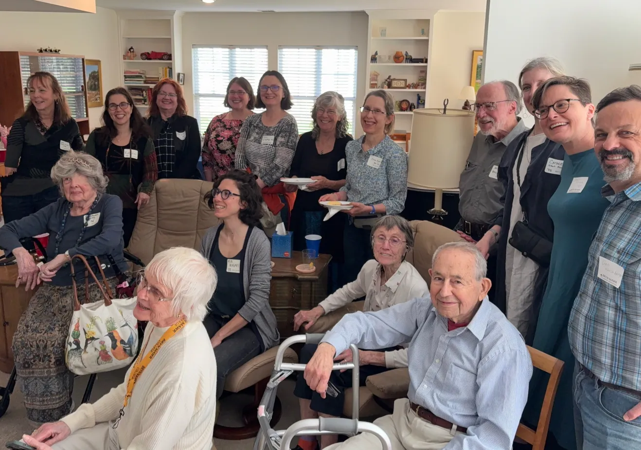 A group of older men and women stand and sit in a room.