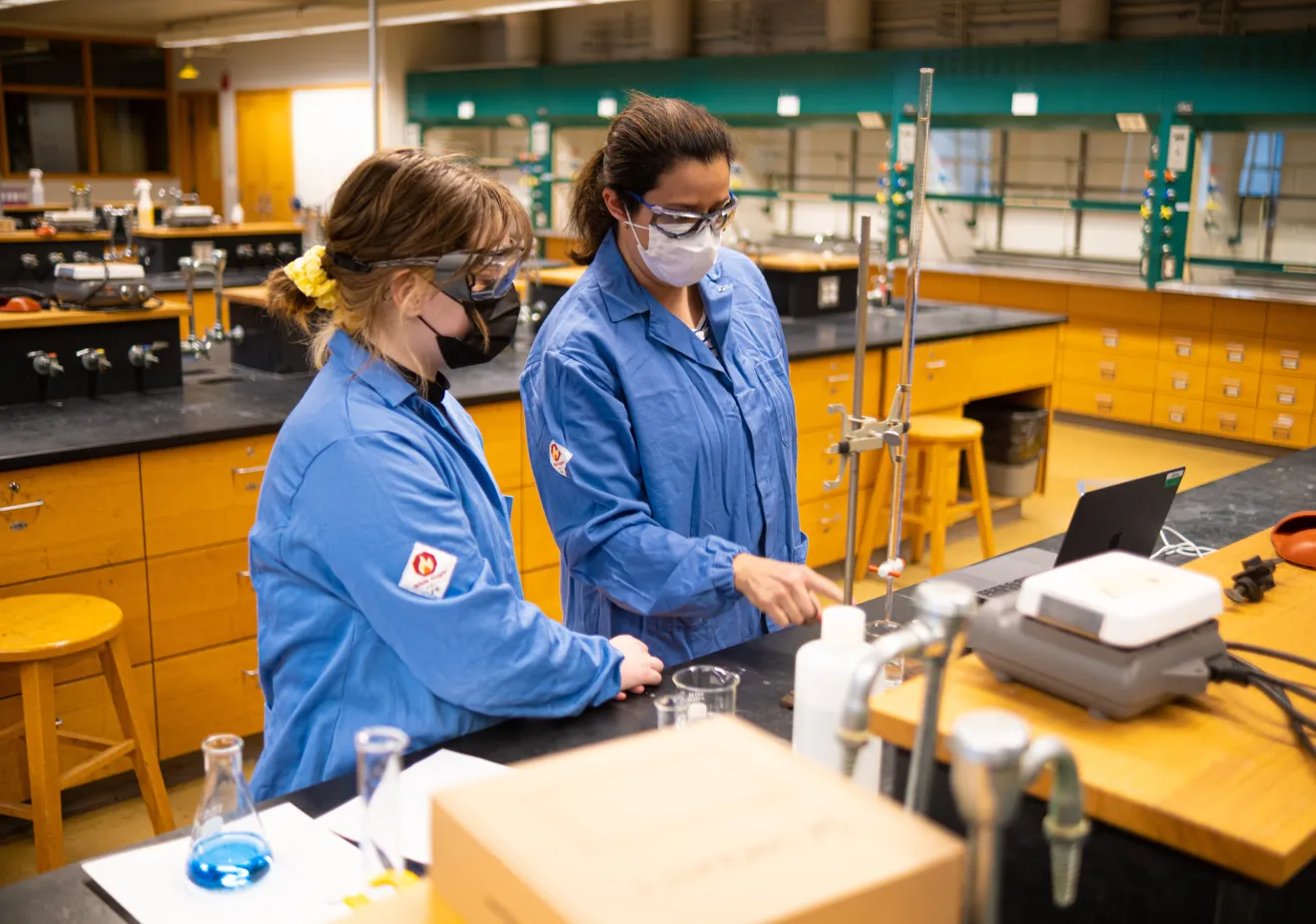 Two women in protective gear work with a long glass tube in a lab
