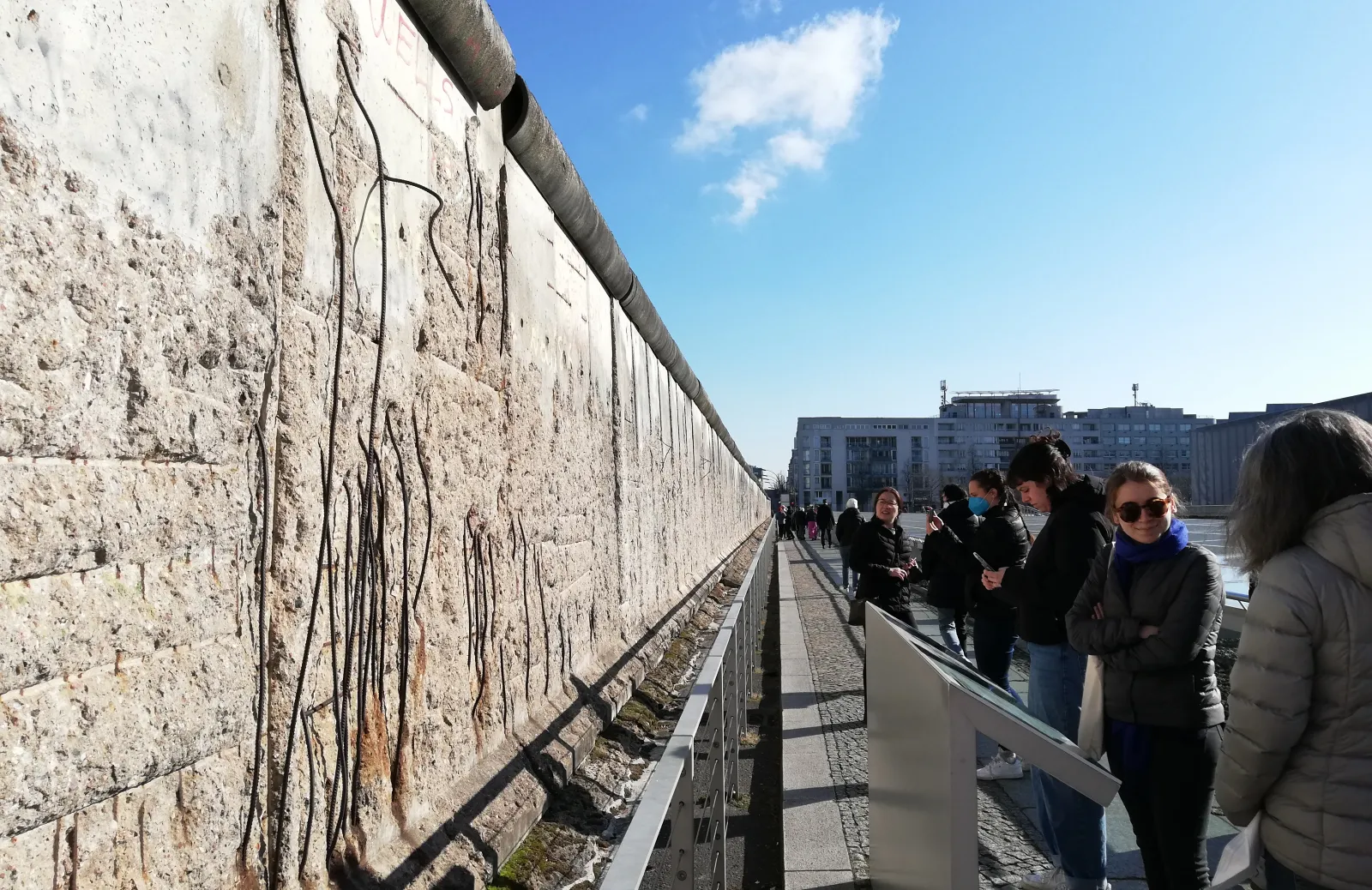 A stone wall with line drawings of figures on the left side of the frame; a row of 6 students and faculty on the right side of the frame observe and read a plaque about the art under a blue sky.