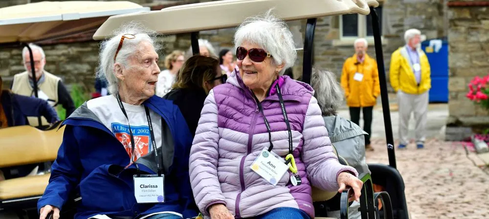 Two older women sit and chat on the back of a golf cart.
