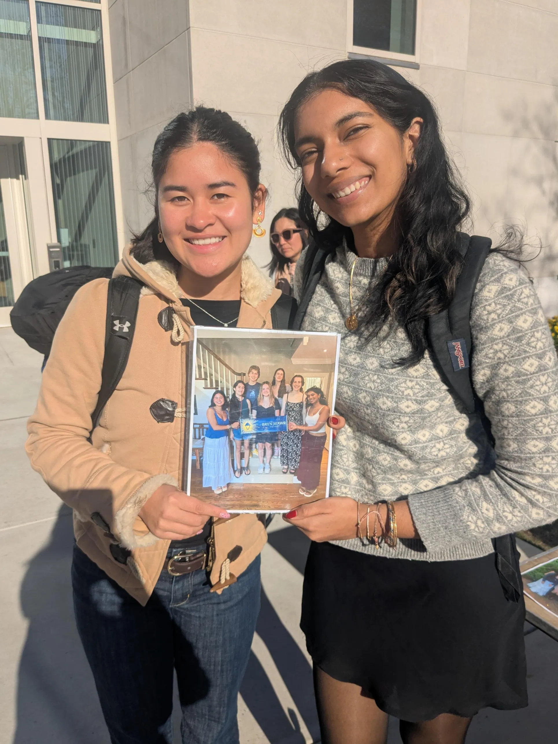 Two students stand out the Well, holding a photo of when they met at a Send-Off Party.