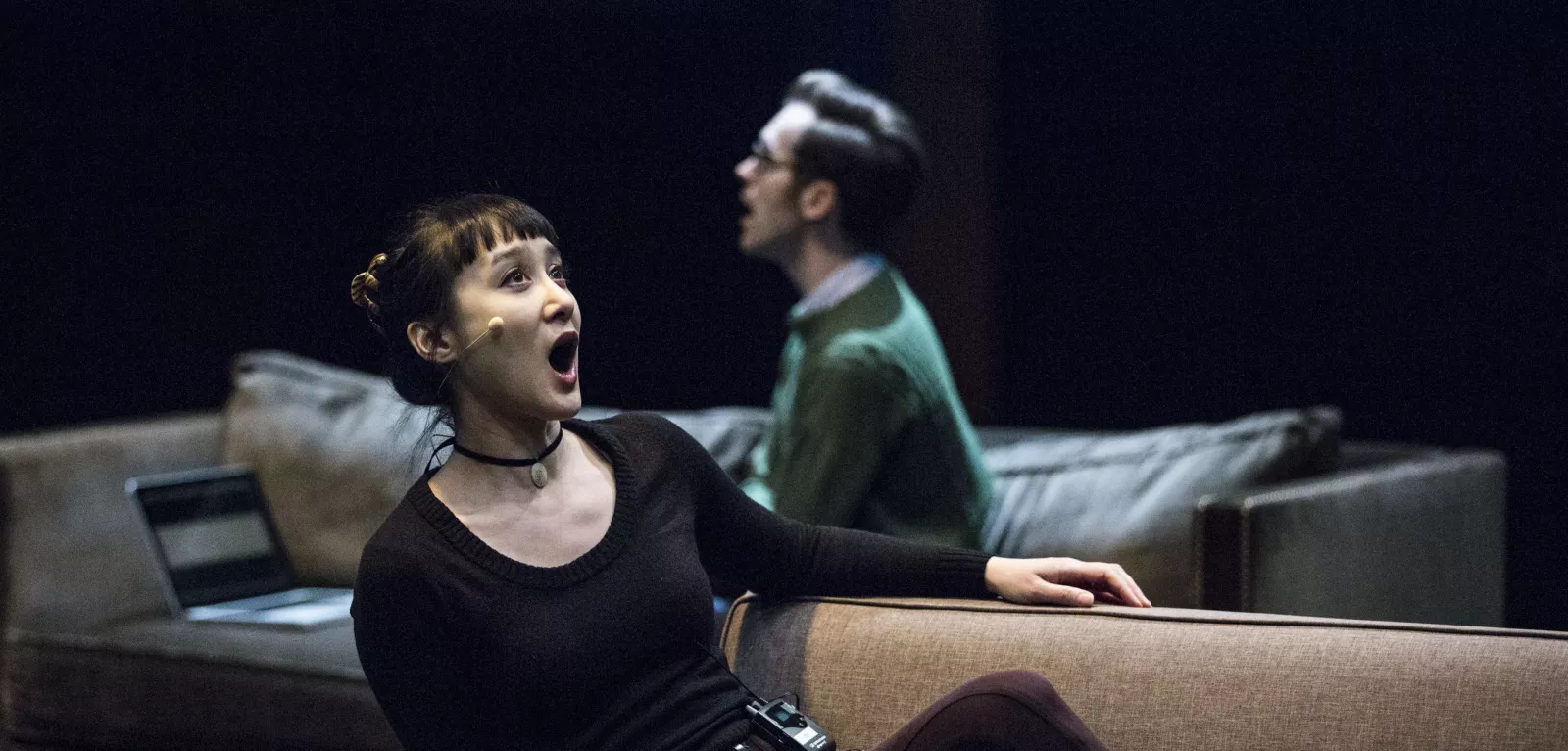 A woman in the foreground sits on a couch facing right and sings. A man in the background on a couch faces left.