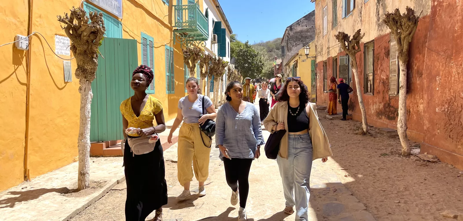 Four students walk down a dirt road with brightly colored two-storey buildings on either side.