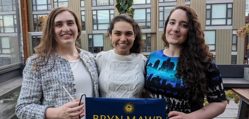 Three young alums smile at the camera. The are on the balcony of a building.