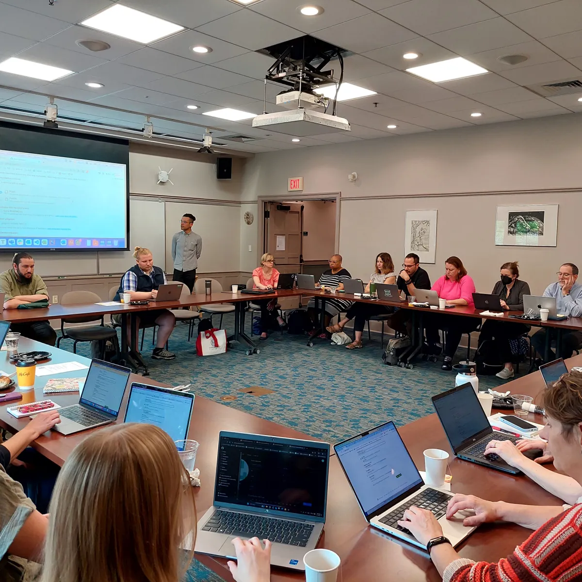 A room full of people with laptops listening to a speaker