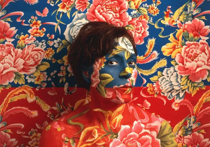 Self Portrait of Artist wearing blue and red floral make-up as camouflage with wallpaper behind her
