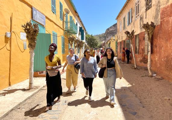 Four students walk down a dirt road with brightly colored two-storey buildings on either side.