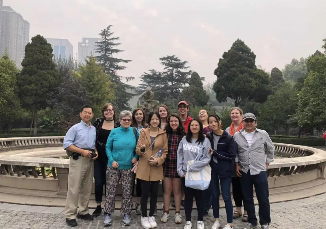 Students and faculty gather in two rows in front of a fountain, with green trees behind them.