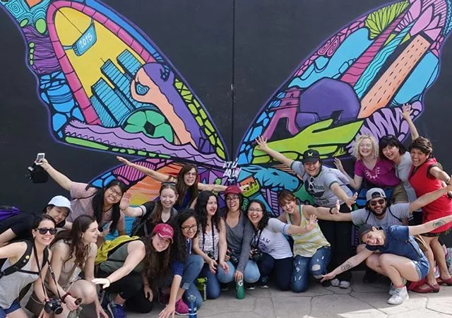 Students pose in front of a butterfly mural, with arms outstretched like wings.