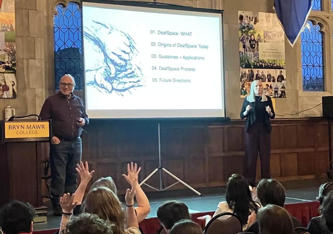 Hansel Bauman presents about DeafSpace on stage at the Great Hall. A presentation behind him shows an illustration of hands signing.