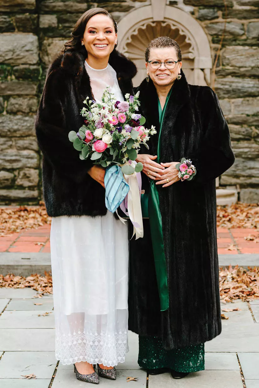 Two people standing in front of a stone wall, one holding flowers.