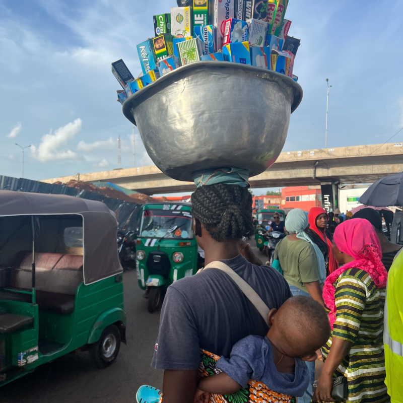 Woman carrying a bowl of goods on her head and a baby on her back. 