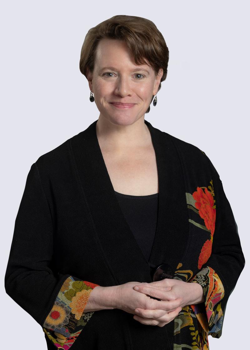 A woman with short, brown hair smiles at the camera. She is wearing a black shirt and a black blazer with embroidered cuffs.