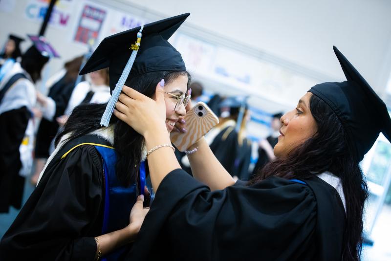 Two 2024 graduates smiling in their regalia. One is adjusted the other's cap.