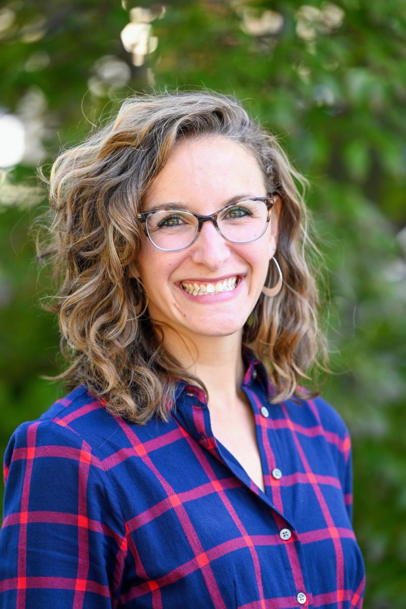 A woman with blond wavy hair smiles at the camera. She is standing in front of greenery. She is wearing a blue and red plaid shirt, glasses, and silver hoop earrings.